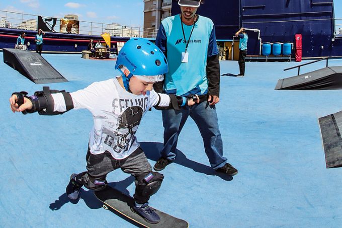 Menino aprende a andar de skate em pista azul. Ele está acompanhado de um monitor