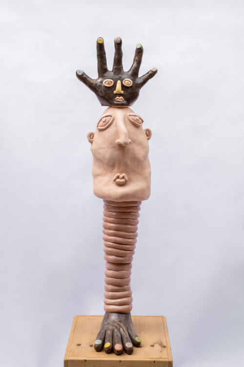 Escultura em cerâmica em forma de totem com um pé negro na base, um pilar anelado, um rosto branco e uma mão negra no topo
