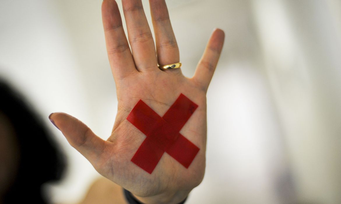 Foto mostra palma de uma mão aberta com adesivo vermelho em formato de X