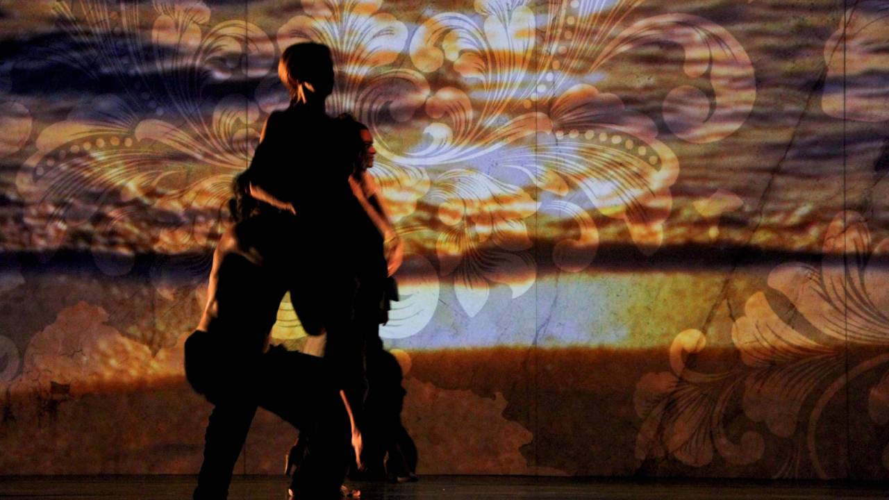 Imagem mostra dois bailarinos dançando em palco, em frente a projeção de entardecer