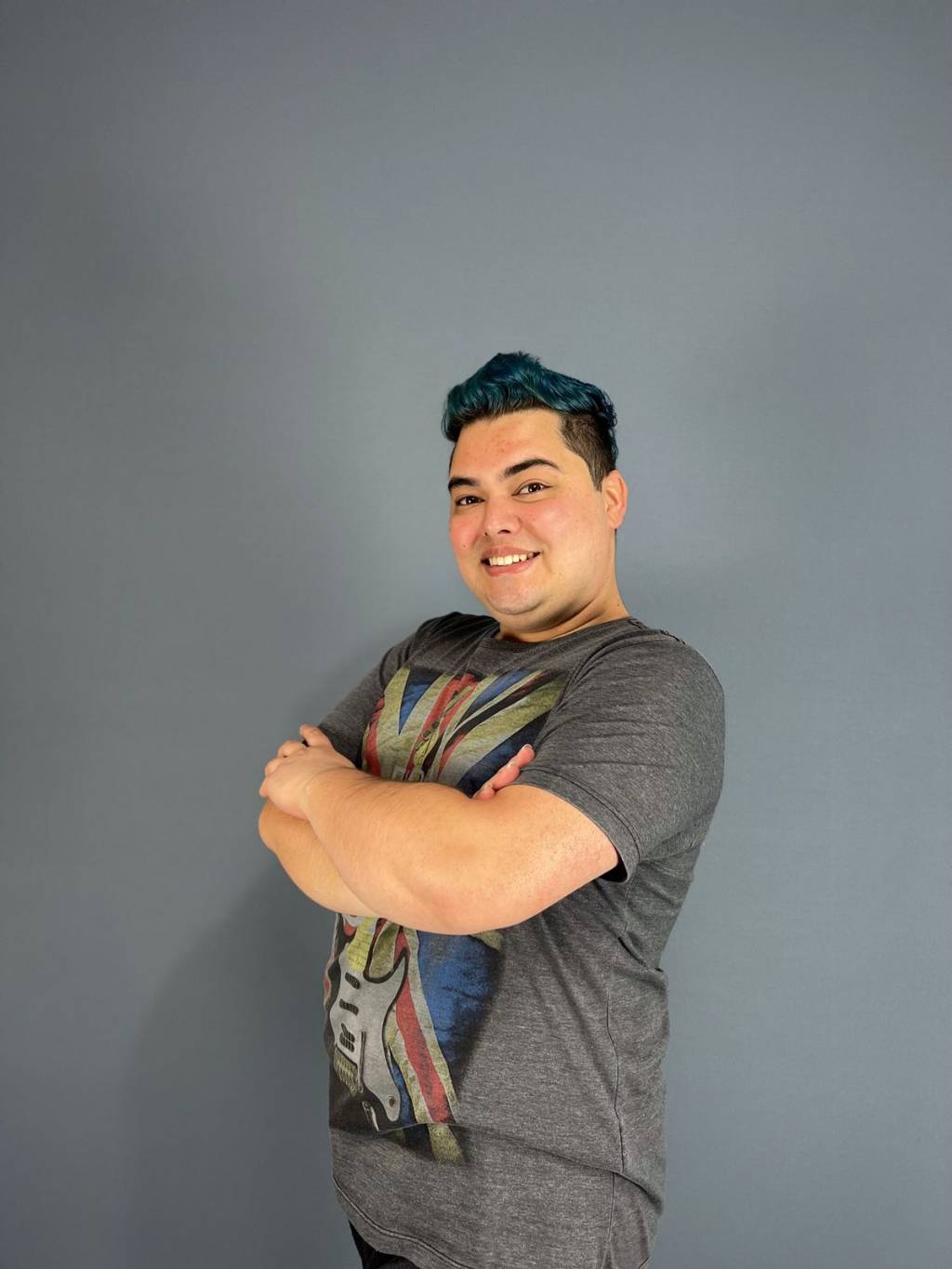 Bruno Fuziwara (David Costa-Divulgação) Homem branco de cabelos pretos sorri de braços cruzados e veste camiseta cinza.