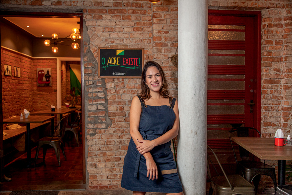 Amanda Vasconcelos posa ao lado de pilar branco à frente de parede de tijolinhos e placa onde se lê Casa Tucupi