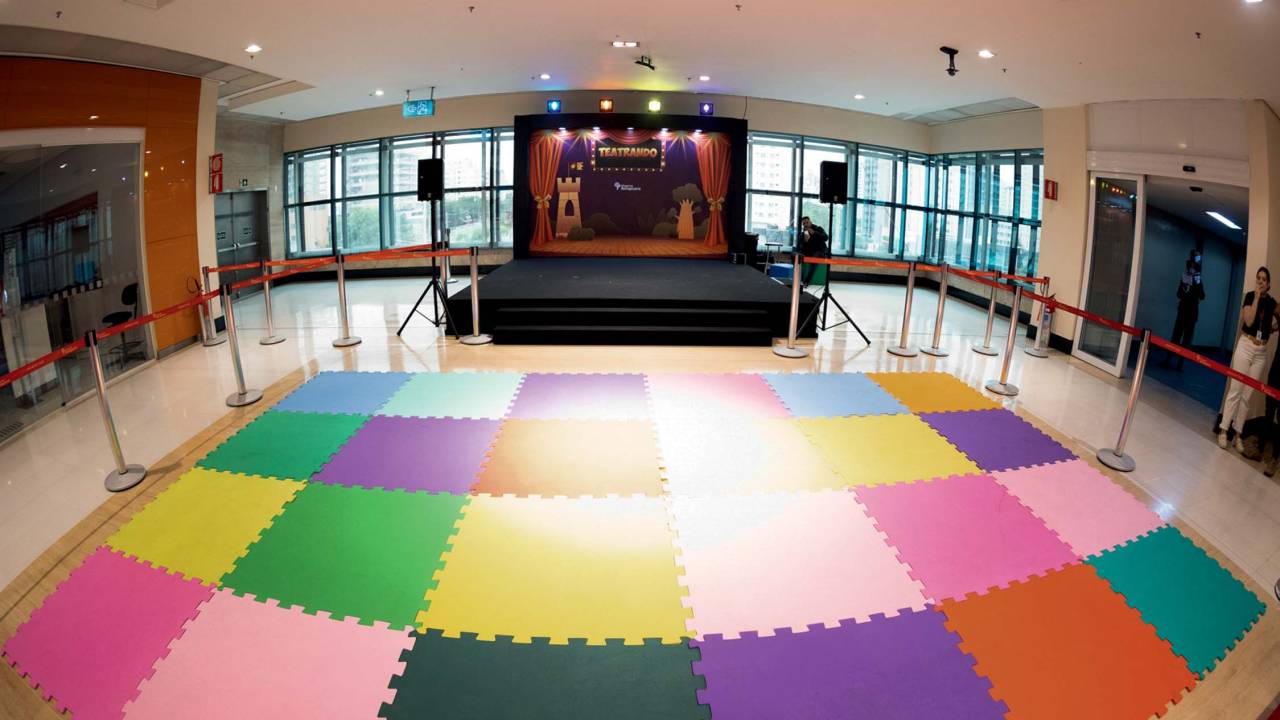 Palco de teatro em um piso do Shopping Ibirapuera. No chão está um tapete com quadrados coloridos para as crianças sentarem