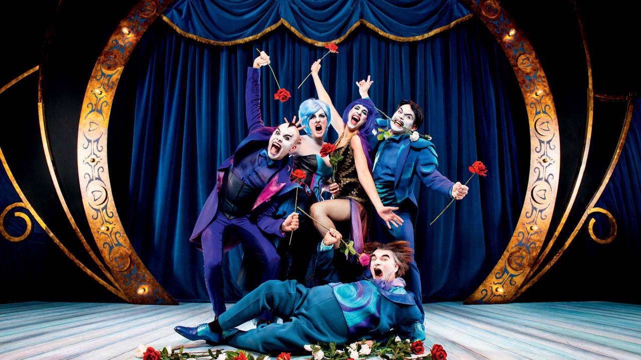 Cantores-atores de espetáculo humorístico de ópera. Usam figurinos extravagantes azuis e maquiagens caricatas. Posam em um palco iluminado, com cortinas azuis e alguns seguram rosas na boca. São três homens brancos e duas mulheres brancas, que usam perucas azuis