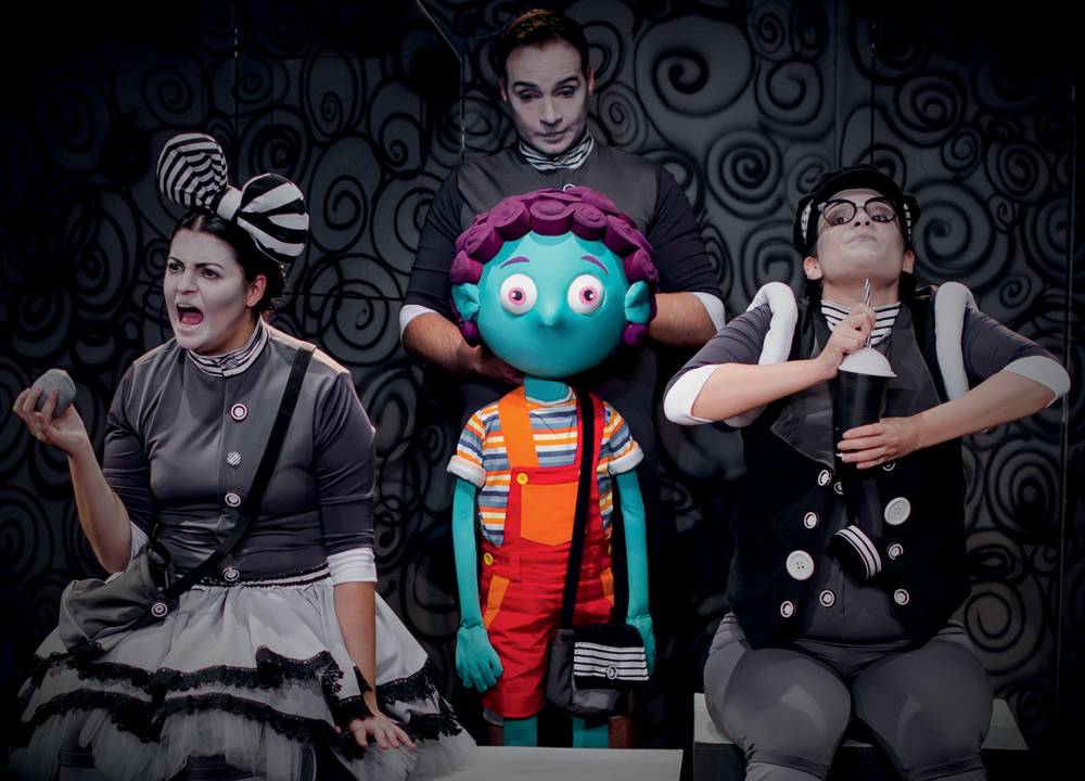 Três atores vestem roupas em preto e branco. Eles interagem com uma marionete de um menino todo colorido