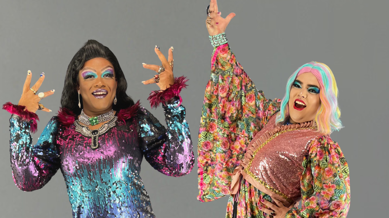 Duas drag queens estendem as mãos e posam sorridentes. A da esquerda é negra e veste vestido nas cores roxo, azul e rosa. A da direita é branca, com peruca branca e azulada, e veste peça florida.