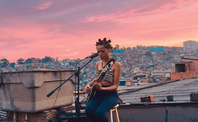 Imagem mostra mulher tocando guitarra sentada em banco, com horizonte urbano ao fundo
