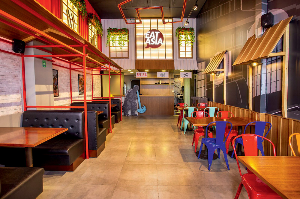 Ambiente interno do restaurante com coberturas remetendo a estabelecimentos e casinhas, paineis com referências ao Godzilla, mesas e cadeiras