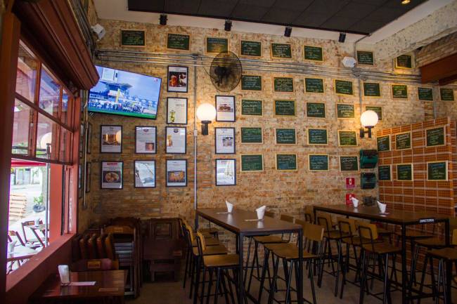 Ambiente interno de bar com tijolinhos à vista e quadros na parede, mesas e cadeiras e um telão de televisão no canto