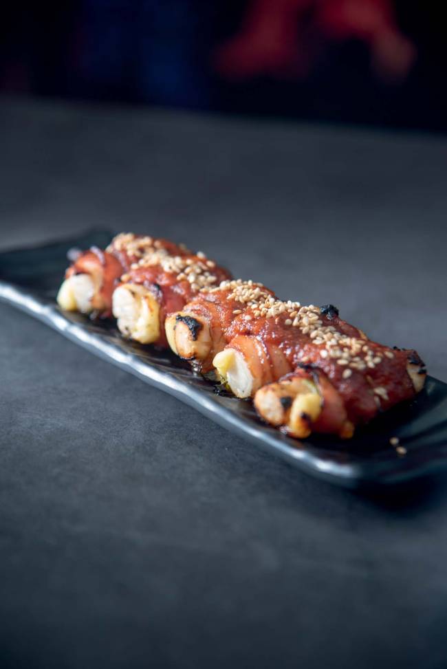 Bacon ddeokkochi (espetinho) – Izakaya – The Oriental Izakaya Espetinho de massa de arroz com bacon sobre prato comprido de cerâmica preta