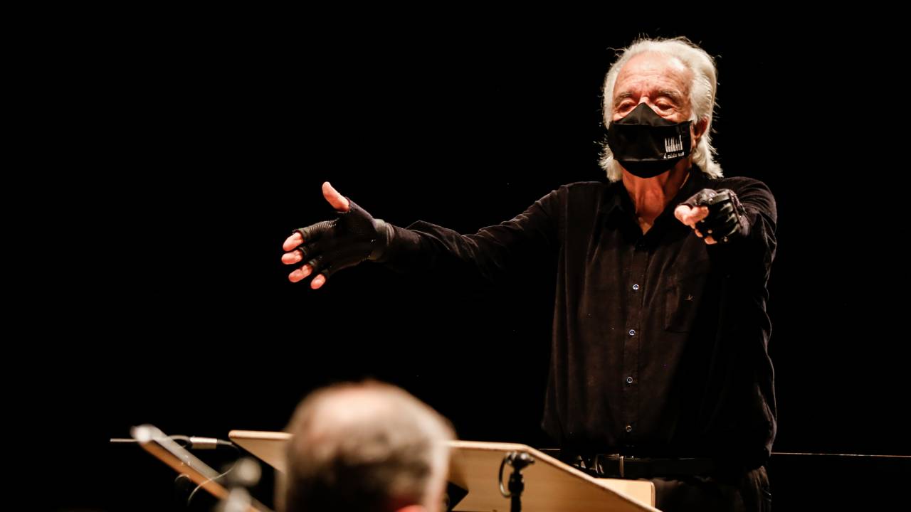 Imagem mostra maestro grisalho, com máscara e luva pretas, regendo orquestra