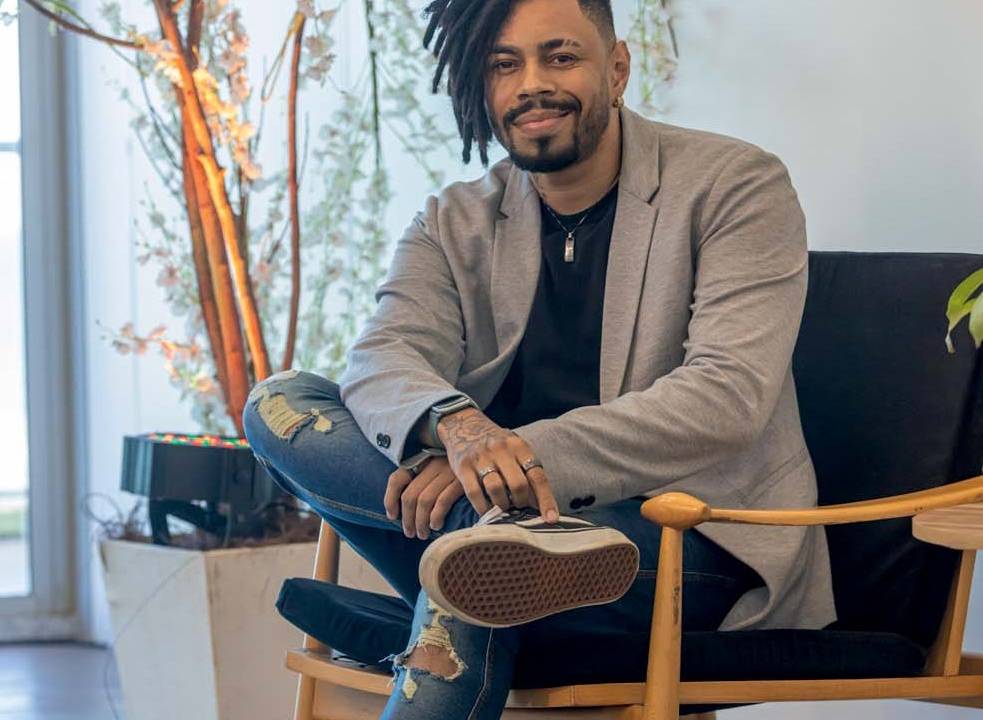 Ale Santos, um homem negro, usando terno e calça jeans, sentado em uma cadeira sorrindo