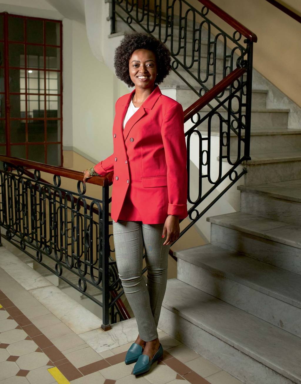 Secretária de Cultura da cidade de SP Aline Torres posa apoiada no corrimão de uma escada. Ela é uma mulher negra, de cabelos crespos e escuros estilo black power. Usa uma calça cinza e um blazer rosa e sorri para a câmera