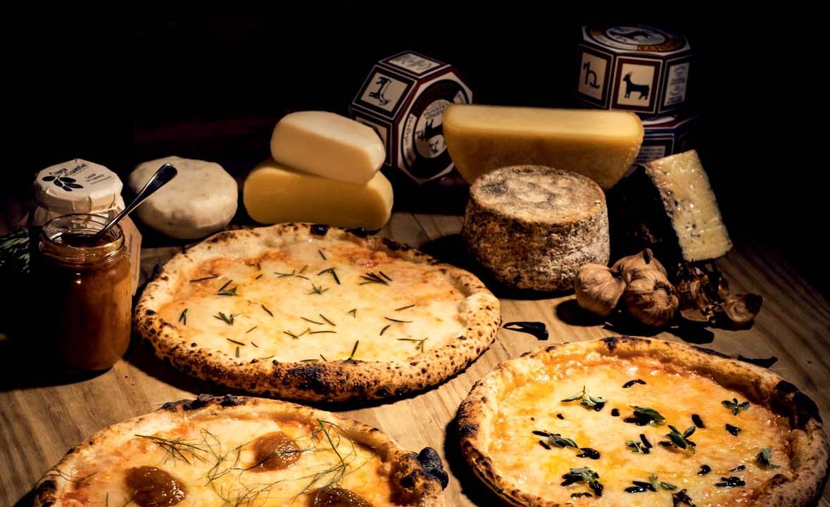 Três pizzas sobre mesa de madeira rústica ao lado de diversos cortes de queijo