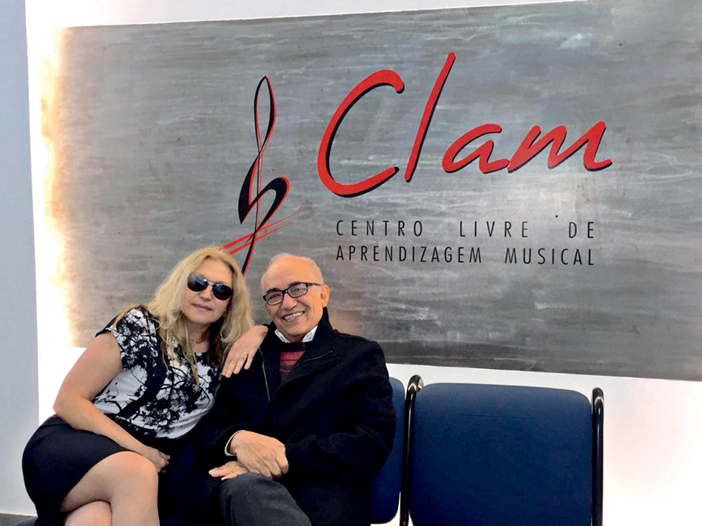 Clam 50 Anos Imagem mostra mulher loira sentada ao lado de homem careca, em sofá