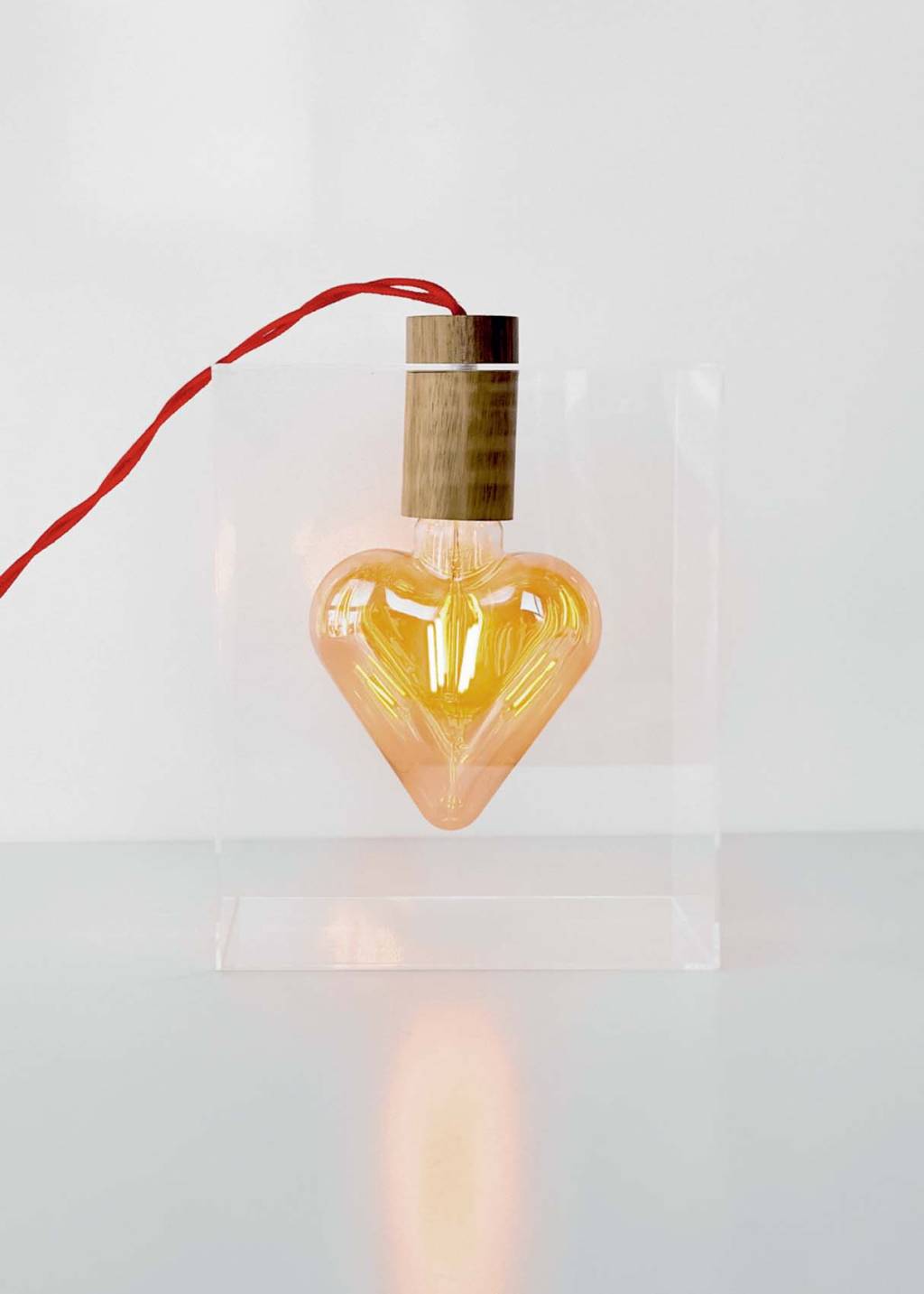 Luminária em formato de coração com bocal em madeira. O coração está posicionado dentro de um quadrado transparente, enquanto seu fio vermelho sai para fora