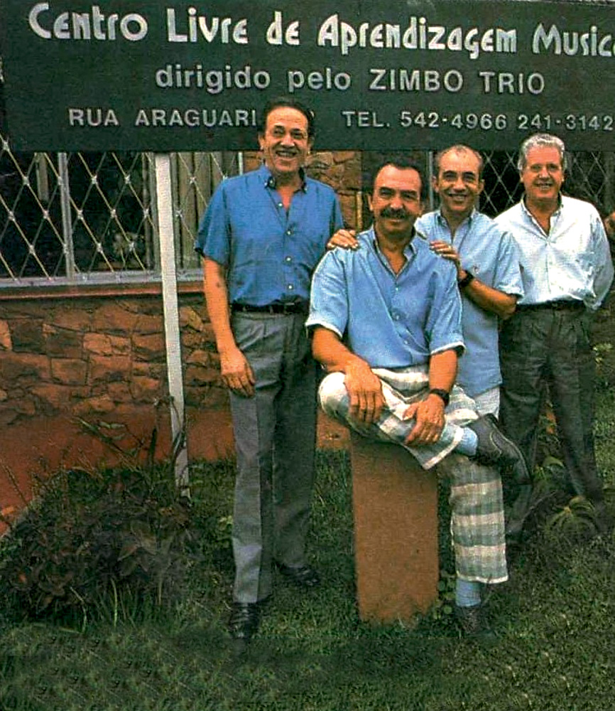 Imagem mostra quatro homens de camisa social sorrindo, ao lado de placa de escola