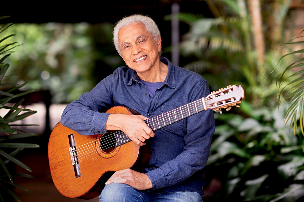 Imagem mostra homem grisalho sorrindo, segurando violão no colo