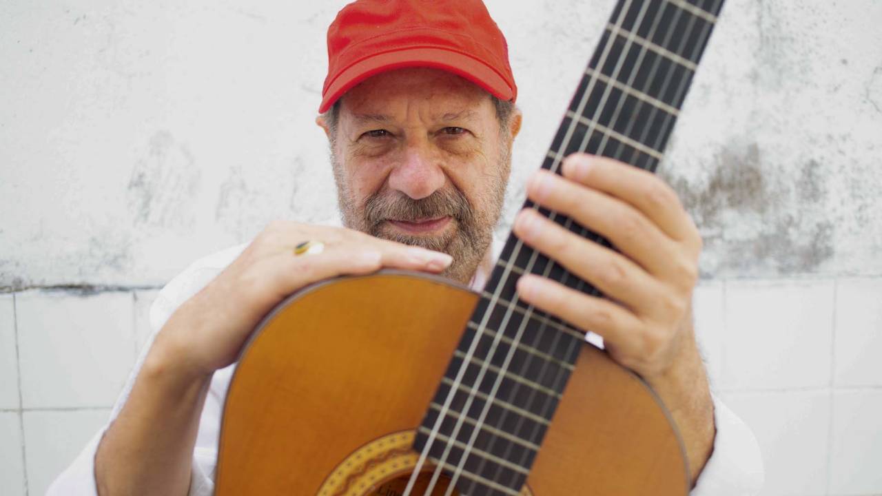 Imagem mostra homem de boné vermelho segurando violão.