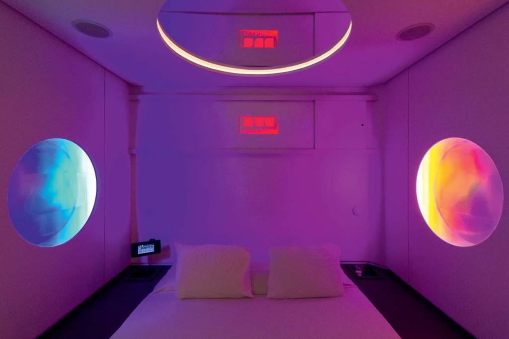 Foto mostra cama em quarto de motel com luz LED em tom roxo pastel