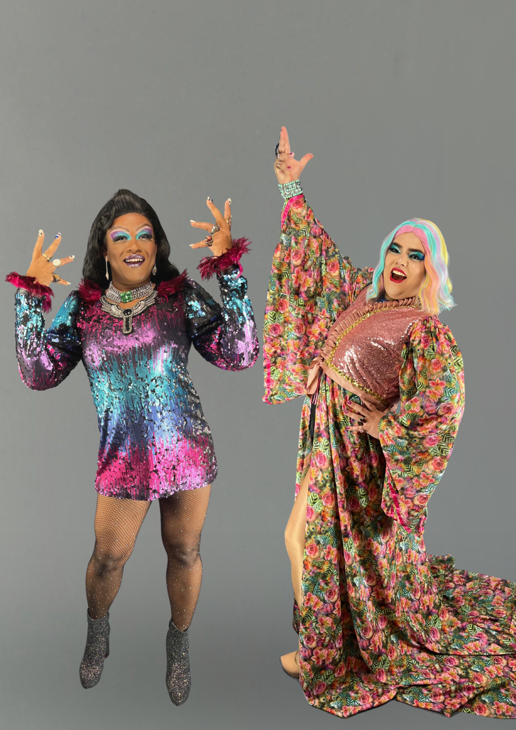Duas drag queens estendem as mãos e posam sorridentes. A da esquerda é negra e veste vestido nas cores roxo, azul e rosa. A da direita é branca, com peruca branca e azulada, e veste peça florida.