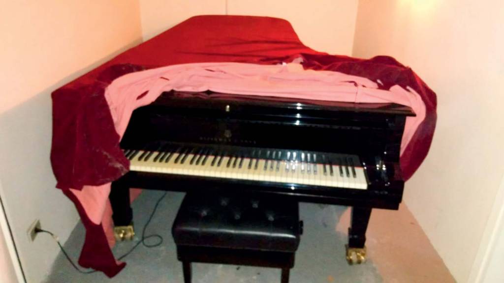 Piano Steinway & Sons antigo, coberto por um tecido de veludo vermelho. Está em um cômodo do hotel Maksoud Plaza que tem piso de carpete cinza