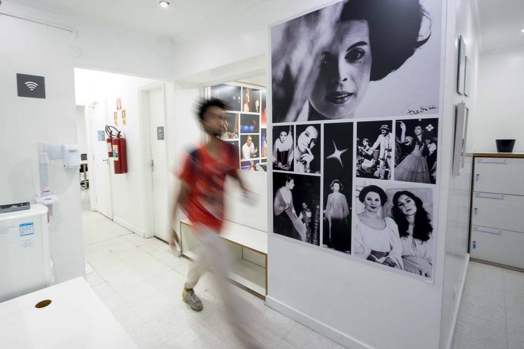 Foto mostra estudante de camiseta vermelha passando por corredor branco com retratos em preto e branco na parede.