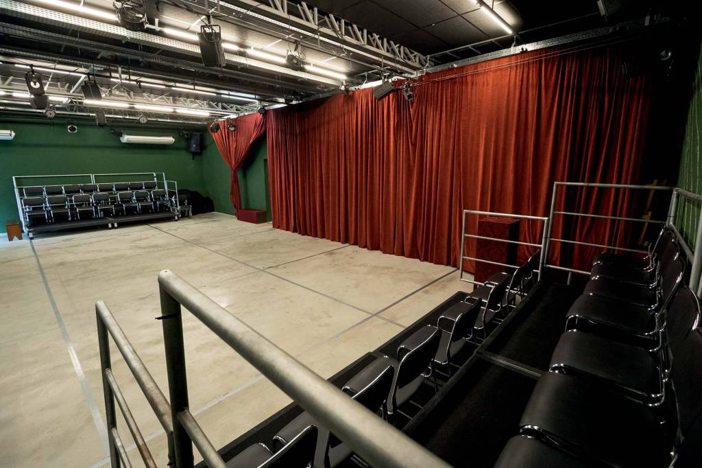 Foto exibe palco com arquibancadas pretas e cortina vermelha.
