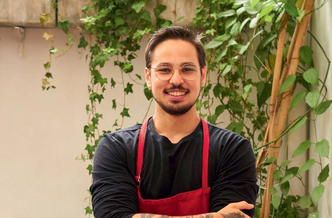 Homem de camisa preta vestindo avental vermelho e com os braços cruzados em frente a parede bege e tendo plantas ao fundo