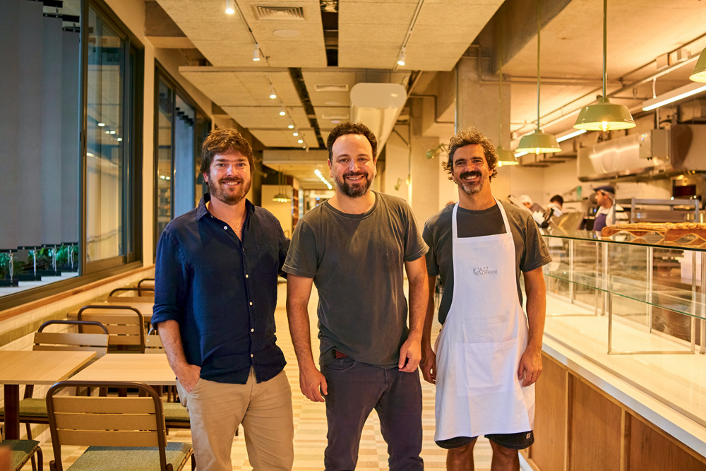 Três homens dentro de ambiente onde há mesas e iluminação amarela