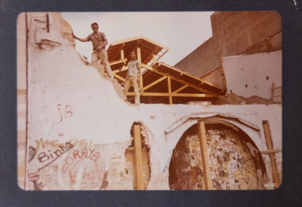 Imagem mostra construção de prédio com dois homens em cima de parede