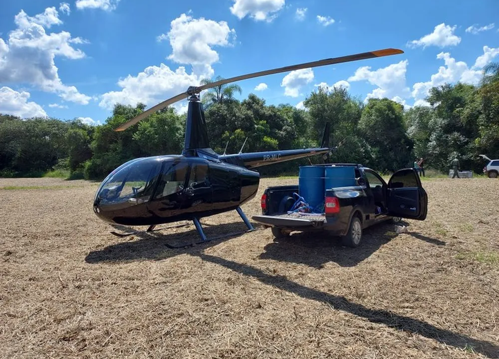 Foto exibe helicóptero preto e pickup preta ao lado em terreno.