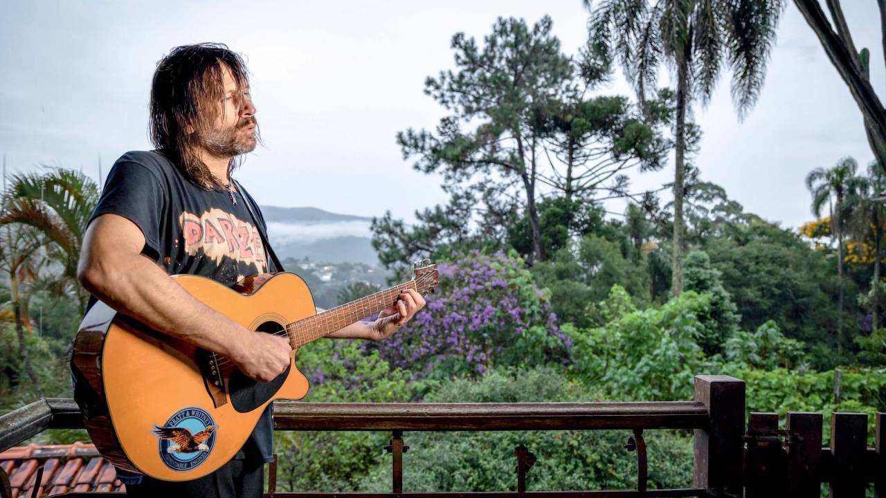 Imagem mostra homem tocando violão, com os cabelos molhados, em pé, em uma sacada com uma paisagem de serra ao fundo.