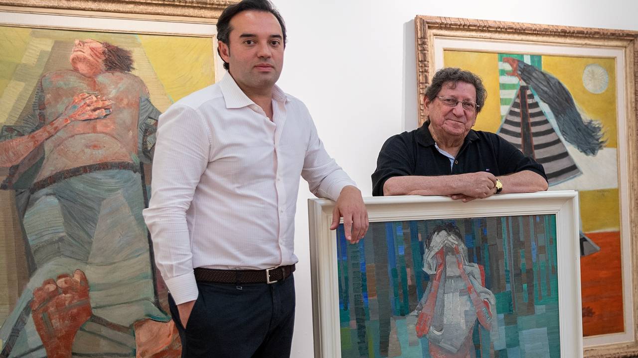 Acacio Lisboa, responsável pela Galeria Frente, e o curador Jacob posam lado a lado com quadro em tons verdes de pé na frente.