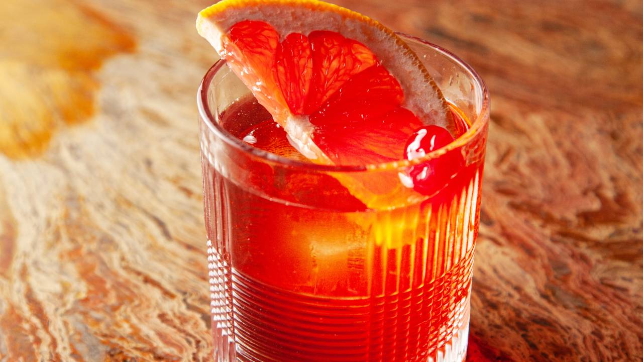 Drink alaranjado em copo baixo sobre mesa de madeira com meia fatia de laranja dentro