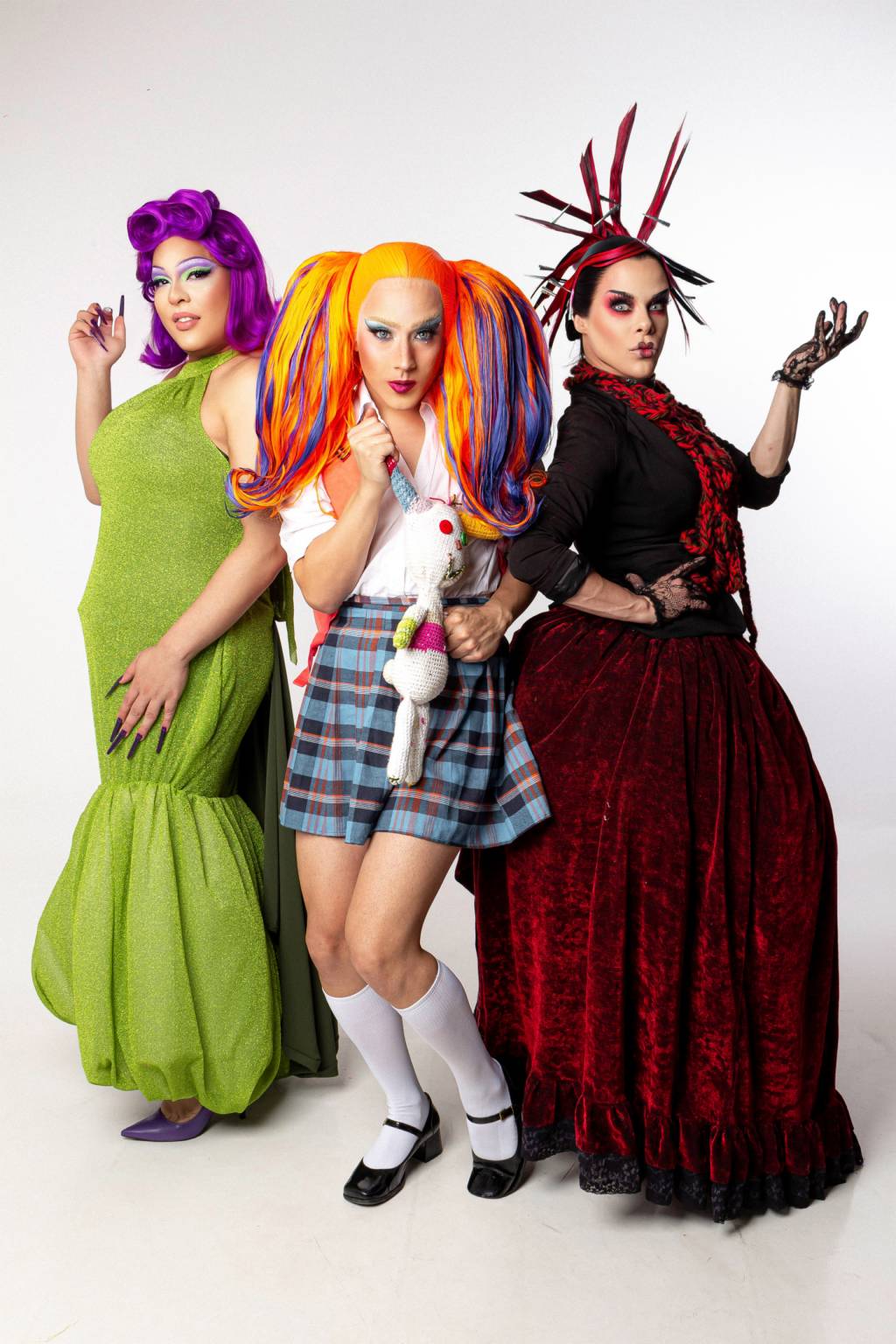Três drag queens posam de pé em fundo branco. A partir da esquerda, vestem vestido verde, roupa de menina colegial e vestido vermelho escuro de vilã.