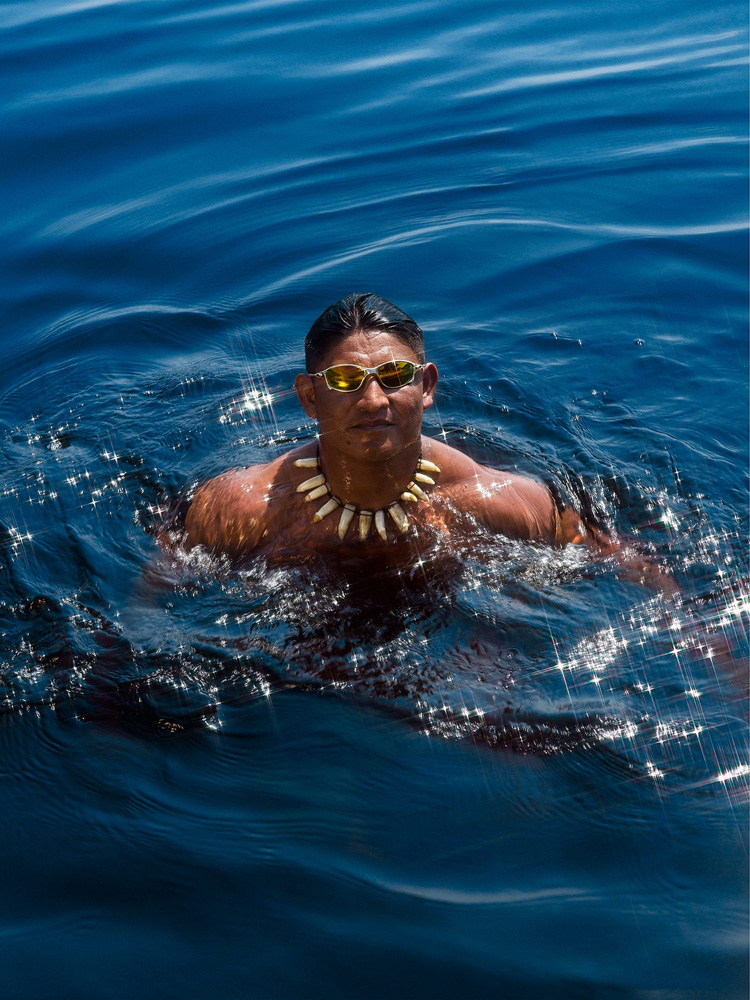 Imagem mostra homem indígena em um rio, usando óculos escuros
