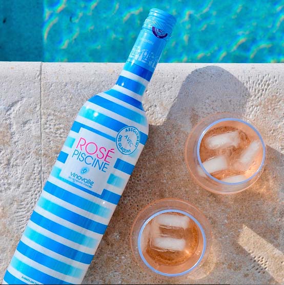 Garrafa de rosé listrada de azul e branco com dois copos ao lado contendo vinho e cubos de gelo, na beirada de piscina