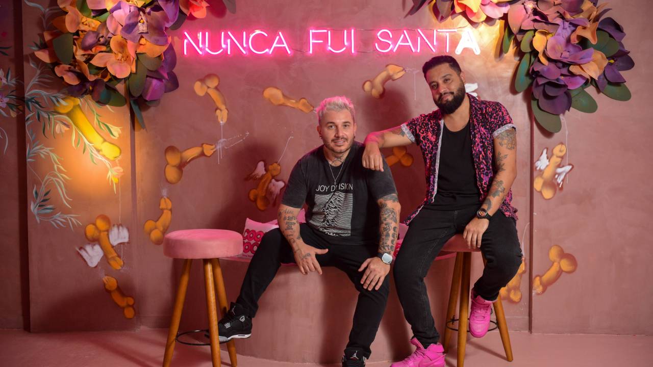 Dois homens posam sentados em local com fundo rosado e neon com a frase NUNCA FUI SANTA.