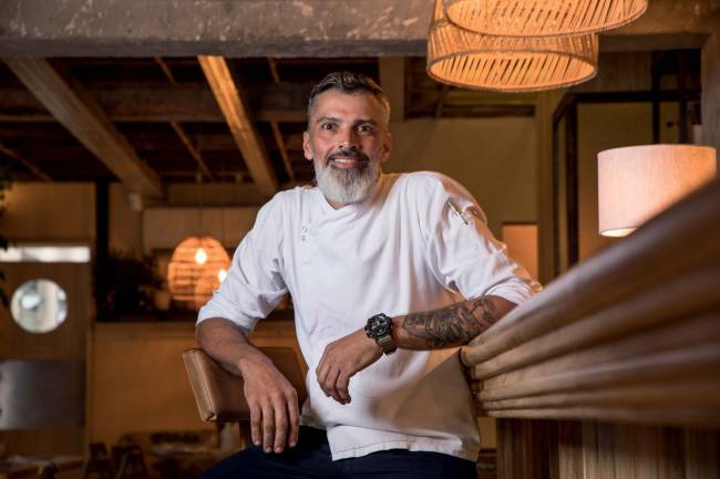 Chef Kleber Klock Azay Homem de barba grisalha e cabelo preto sentado em cadeira com os braços apoiados nas costas da cadeira e na mesa usando uma roupa branca