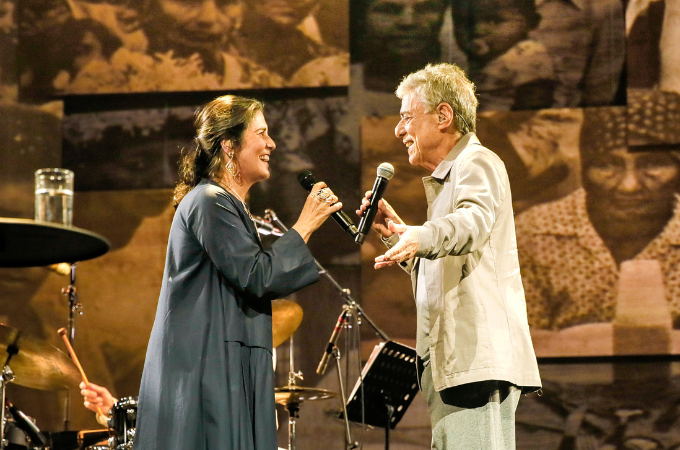 Imagem mostra mulher e homem segurando microfone, ambos sorrindo, em cima de palco
