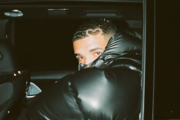 Drake está usando uma jaqueta puffer preta, dentro de um automóvel, exibindo parte de seu rosto