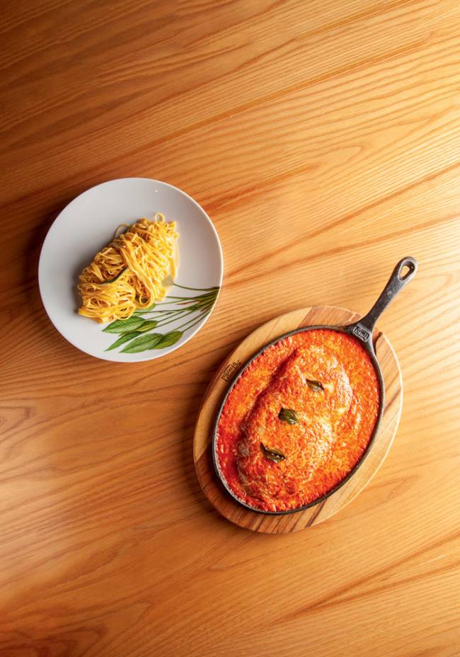 Parmigiana em panela de ferro sobre tábua ao lado de prato de porcelana com tagliolini na manteiga de sálvia