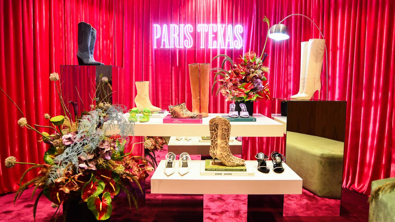 Neon rosa 'Paris Texas' aparece em destaque em frente a cortina de veludo e ilumina algumas bancadas claras e repletas de sapatos e botas.