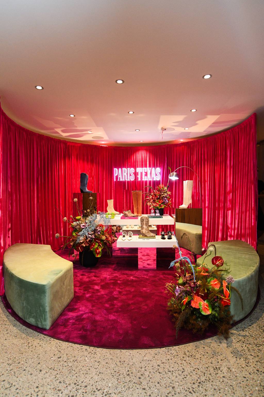 Neon rosa 'Paris Texas' aparece em destaque em frente a cortina de veludo e ilumina algumas bancadas claras e repletas de sapatos e botas.