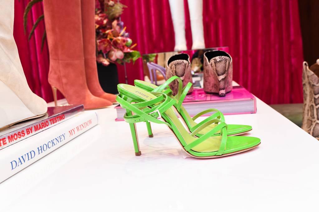 Neon rosa 'Paris Texas' aparece em destaque em frente a cortina de veludo e ilumina alguns pares de sapato, especialmente um par verde em destaque.