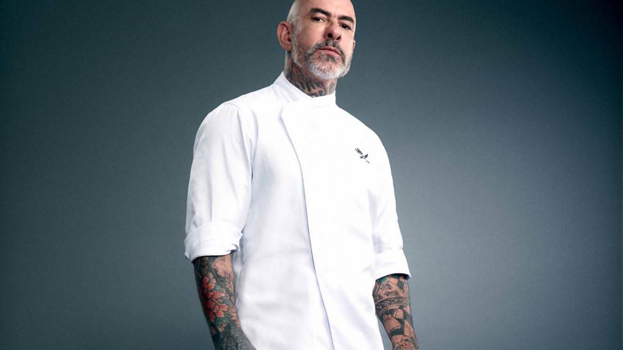 Foto de Henrique Fogaça, com fundo neutro, semblante sério, usando camiseta de cozinheiro