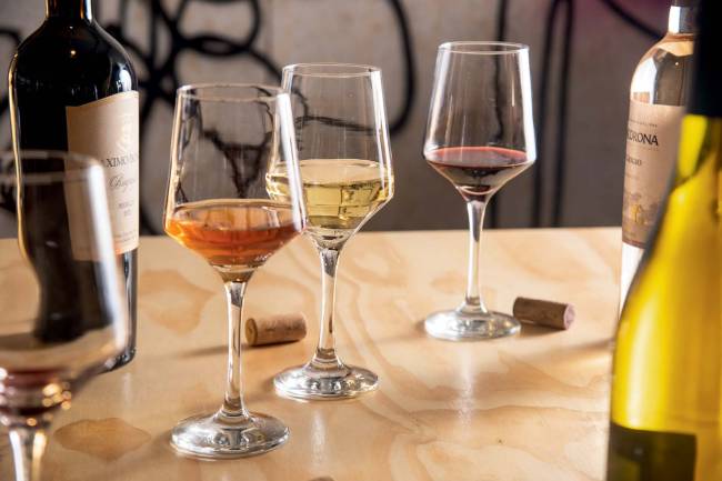 Três taças contendo vinhos branco, rosé e tinto sobre mesa de madeira clara