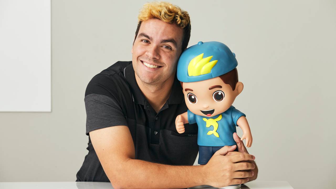 Foto mostra Luccas Neto usando camiseta preta abraçado a um boneco seu, que está em uma mesa, sorrindo