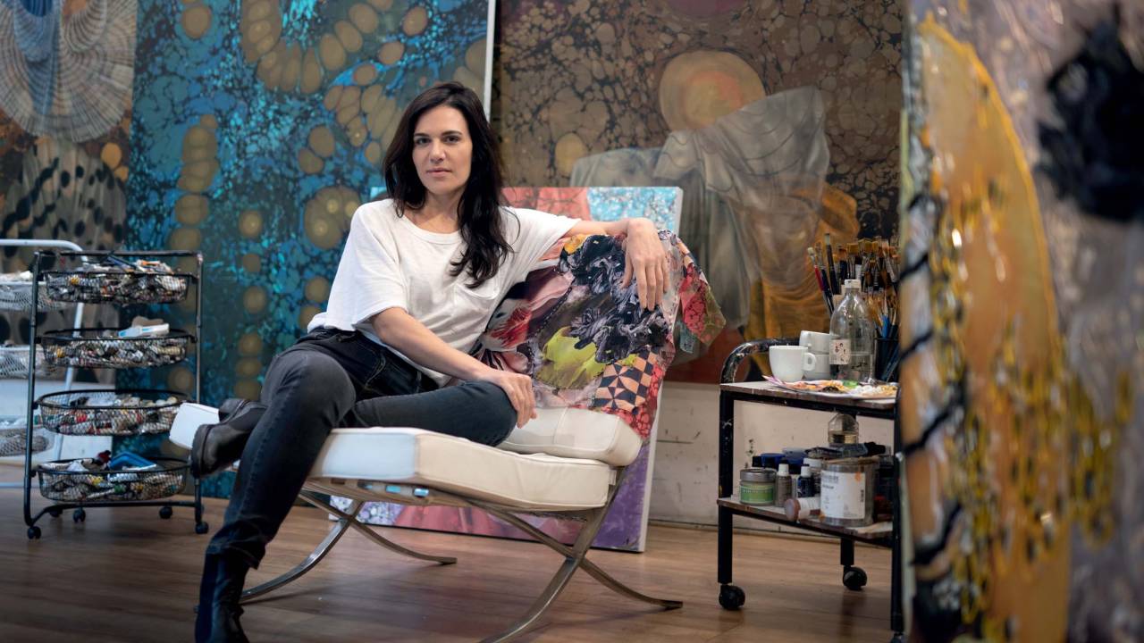 Foto de Mariana Palma sentada, sorrindo, usando calça jeans e camiseta branca, em ateliê